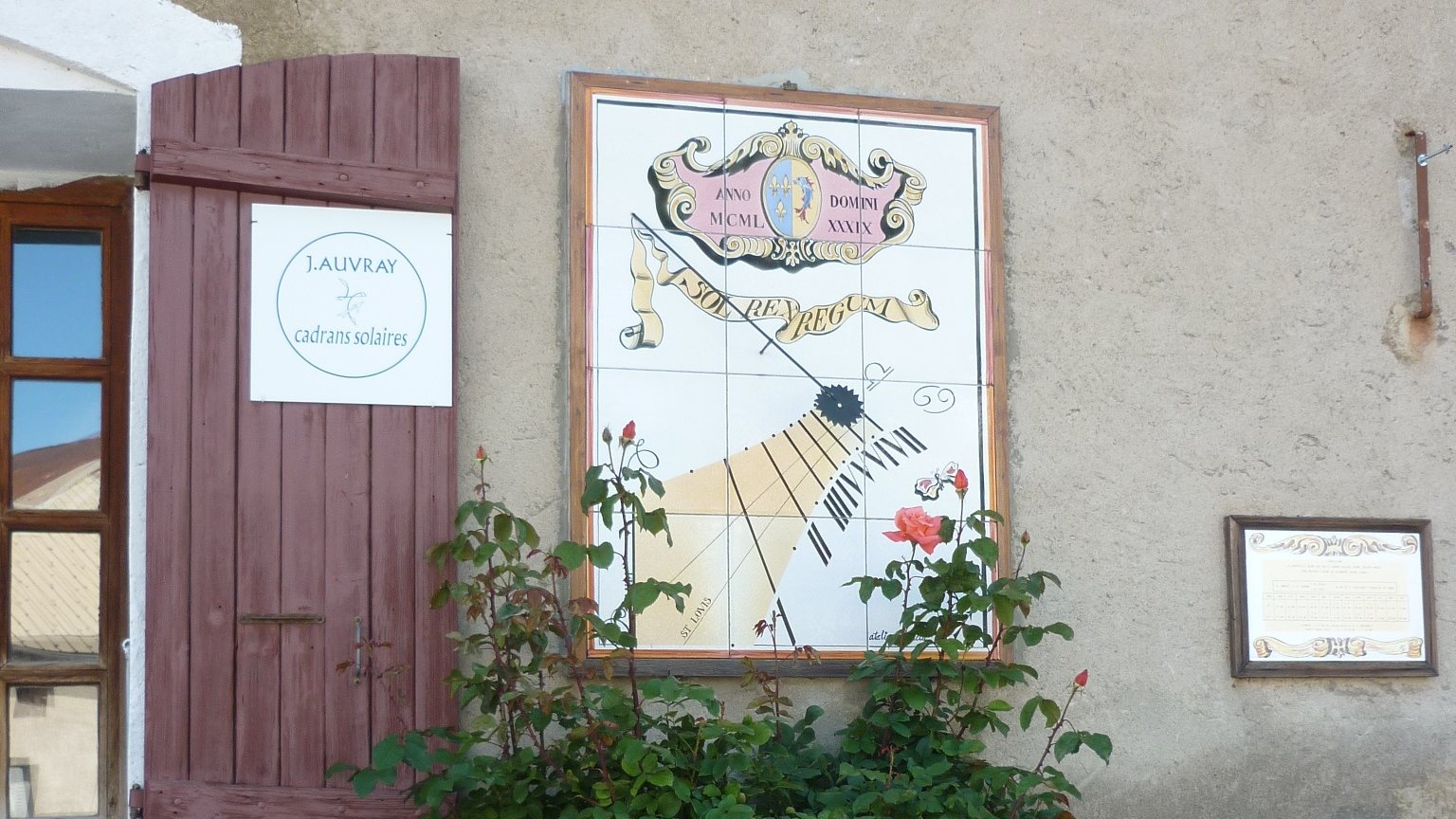 Façade de l'Atelier Acacia à Mont-Dauphin : cadran solaire en carreaux émaillés, volet rouge portant la plaque « J. Auvray cadrans solaires », rosier en fleurs.
